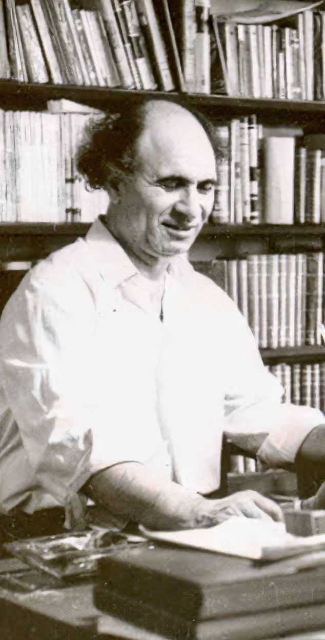 Master Jalil Ziapour in a white shirt with rolled-up sleeves, smiling at his desk with an open book, in front of shelves full of books
